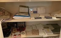 Two shelves filled with linens including folded sheets in original packaging, flat folded linens, Christmas themed cloths, and some boxed items on bottom shelf.