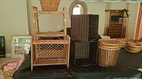 Wicker floor shelf, two square wicker storage boxes with handles, black and dark brown, two round wicker waste baskets, and smaller wicker wall shelf in the background, all placed on a table.