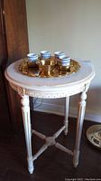 Round white painted wood side table with carved legs and X cross stretcher visible, holding a decorative gold tray with four small demitasse cups and gold sugar bowl on top.