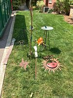 Wide photo showing all lot items arranged on grass including metal sun decorations, ceramic turtle, metal sundial, metal plant supports, bamboo sticks, spinning sprinkler, and hummingbird feeder