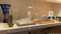 Shelf display showing blue cut-to-clear glass vase, other clear glass vases, brass vases, trays, and boxed candles.