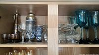 Shelf with various glassware including decorated decanter, stemmed glasses, clear etched goblets, and blue-green hand-blown wine glasses