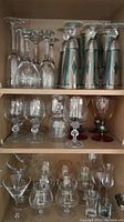 Shelf with various vintage stemware; clear wine glasses, champagne flutes with green and gold design, cut glass goblets, and colored stemware.