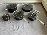 Top-down view of the five Heritage The Rock pots and pans arranged with their lids on a grey sheet.