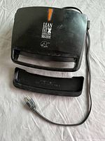 Top view of closed black George Foreman Grill with power cord and drip tray detached, showing external condition and branding.