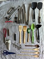 Photo of assorted kitchen utensils laid on fabric including tongs, spatulas, whisks, garlic press, can opener, potato mashers, ladles, wooden spoons, scissors and metal hooks.