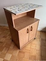 Wooden microwave cart with top floral wallpaper, middle open shelf, two-door cabinet, and casters on wooden floor.