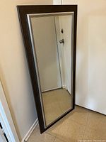 Tall rectangular mirror leaning against a wall, showing clear reflective surface and dark wood frame.