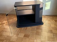 Black rotating TV stand with glass door open showing interior shelf and side drawer on parquet floor in natural light.