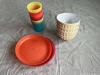 Photo of 4 stacked butter-yellow melamine bowls, 6 stacked multicolor plastic cups, and 2 stacked plastic plates on a light gray fabric surface.