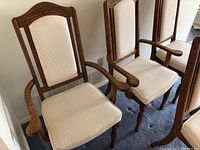 Photo showing three wooden dining chairs with beige upholstered seats and backs. Two are armchairs with wooden arms, one is a side chair. Wood has carved details and medium brown finish. Fabric shows subtle diamond pattern and some discoloration.