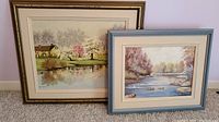 Photo showing two framed landscape art prints leaning against a wall on carpeted floor.
