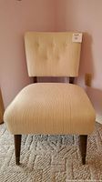 Front view of a cream upholstered accent chair with button tufted backrest and wooden legs against a pink wall.