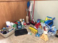 Photo showing multiple cleaning tools and supplies organized on a tiled floor against a wooden backdrop including spray bottles, brushes, cleaning cloths, gloves, dustpan and more.