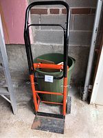 Front view of the orange and black metal moving dolly standing in front of a green plastic garbage can against a cinderblock wall.