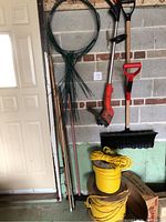 Wide view showing all garden tools including shovel with snow plow blade, orange string trimmer, green stakes, twine, metal rods, and cardboard box