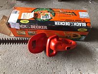 Photo showing the Black & Decker hedge trimmer with 16-inch blade next to its original box, displaying the orange plastic body and serrated blade.