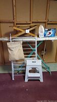 Full view of the lot showing large ironing board with floral cover, electric iron on top, metal drying rack in front, white plastic step stool, and small wooden tabletop ironing board resting on the ironing board.
