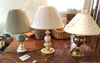 Three lamps placed on a wooden table showing varied bases and shades including a green and gold base, a white decorative base, and a brass base.