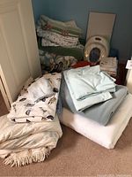 Photo showing a pile of various folded blankets, throws, and linens including a light beige fringed throw and patterned white blanket with leaves.