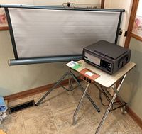 Wide view showing the projector on a foldable table next to the tripod standing projection screen.