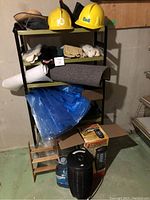 Shelf unit with work gloves, hard hats, insect control box, blue plastic bags, and other miscellaneous items arranged on and in front of the shelving unit.