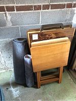 Two sets of wooden folding TV trays/tables with natural wood finish and handles, stacked upright against concrete wall.