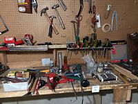 Wide view of workbench with various hand tools laid out such as screwdrivers, hammers, clamps, measuring tapes, utility knives, and small parts containers.