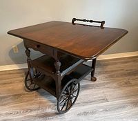Front and side view of the wooden rolling bar cart showing the foldable side extensions extended and large wooden wheels at one side.