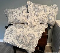 Photo showing queen quilt draped over a chair with two matching pillow shams on top. Quilted fabric is white with gray toile pattern and scalloped edges.