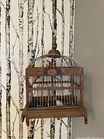 Decorative bird cage hanging or sitting on table against birch bark patterned wallpaper. Shows wood base with floral painted front, metal bars forming cage structure, domed top with finial, two small bird figurines inside.