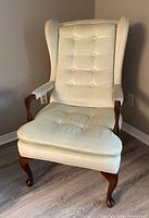 Upright image of a cream-colored wingback armchair with button tufting and wooden legs against a neutral wall and wood floor.