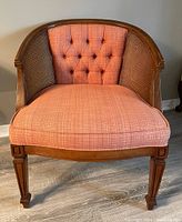 Front view of wood frame bucket chair with salmon pink cushioned seat and tufted backrest, woven cane sides and back, and carved legs.
