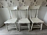 Three white painted wooden spindle back chairs shown from front, highlighting overall condition and paint wear, one chair visibly damaged.