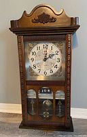 Front view of the wooden pendulum wall clock showing carved top, clock face with Arabic numerals, and visible pendulum behind glass.