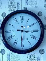 Photo of a large round wall clock with black frame and white face. Clock has Roman numerals and 24-hour markings. Clock hands are visible and indicate a stopped time.