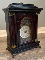 Front angled view of the dark wood table clock showing the ornate gold detailing around the Roman numeral face and the decorative pillars.