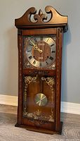 Photo of wooden pendulum wall clock with decorative top, Roman numeral dial, ornate clock hands, and gold-accented glass panel with visible brass pendulum.