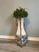 Front view of artificial plant in decorative tall silver-tone metal floor vase on hardwood floor