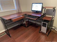 Photo shows L-shaped corner computer desk in a room with hardwood floor and window with blinds. Desk has mahogany finish surfaces supported by black metal frame. Left side surface larger with a drawer beneath. Right side has elevated monitor shelf, sliding keyboard tray and lower shelf holding computer tower case. Monitor turned on displaying a Google search page.