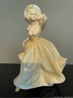 Porcelain figurine of a lady in a flowing pale yellow dress and hat, posed gracefully with hand near head.