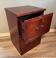 Wooden filing cabinet with two drawers; top drawer is open and has a lock with key on front; the wood has reddish-brown finish; the cabinet is standing on a wood floor next to a wall.