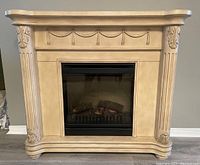 Front view of electric fireplace with decorative cream-colored mantel featuring fluted columns and swag relief detailing. Shows faux logs and embers inside the firebox.