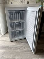 Open view of small white upright freezer showing three wire baskets inside and white sealed door.