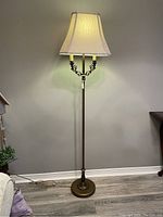 Full view of a metal floor lamp with an ornate stem, two candle-style bulb holders, and a cream fabric shade lit up.