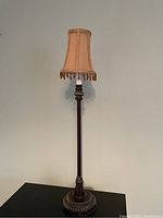 Full view of a tall vintage floor lamp with beige fabric shade and hanging beads, positioned against a plain wall on a dark surface.