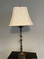 Table lamp on black surface against plain wall, showing full lamp with decorative bronze-tone base, clear spherical accent, and cream fabric shade with floral pattern.