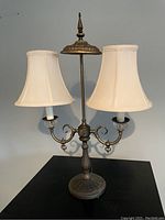 Front view of an antique style metal table lamp with two candle-style sockets and cream fabric shades on black surface against gray wall.
