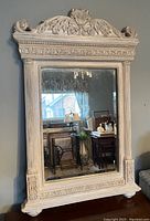 Photo of an ornate, rectangular wall mirror with detailed carved frame hung on a wall, reflecting a room interior with furniture and window.