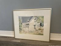 Photo of framed watercolor print depicting a white house with picket fence, standing against a wall.